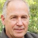 A neatly stacked pile of discarded wooden pallets and packaging materials is situated on a paved driveway next to a warehouse or industrial building. The pallets are of varying sizes, primarily made of rough, unfinished light-colored wood, with some showing visible grain and texture. The materials appear weathered, with surfaces that have minor splinters and signs of wear. The pallets are arranged in an organized manner, with some leaning against the building wall and others lying flat on the ground. In the background, part of a large metal roller shutter door and a section of the brick or concrete exterior wall are visible, suggesting an industrial or commercial environment suitable for private waste collection or site clearance. Natural daylight illuminates the scene evenly, emphasizing the textures and colours of the pallets and surrounding surface. This scene exemplifies a scenario where a rubbish removal service, such as those offered by Big Ben, might facilitate the collection and disposal of bulky, unauthorised waste materials for business or industrial clients, supporting alternative waste handling solutions outside standard local authority services.
