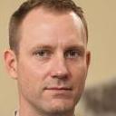 A residential driveway with a large, open-top black skip situated near the front of a modern house with a brick facade and white window frames. The skip is partially filled with mixed waste materials, including blue plastic bags, cardboard boxes, and various debris, visible at the open top. The surface of the skip shows a textured, galvanised steel finish with some rust spots, indicating regular use. To the left of the skip, a pair of orange work gloves rests on the concrete pavement, which is clean and free of loose rubbish. In the background, a wooden fence and a small garden area are visible, with a few green plants and bushes. The scene appears to be captured during daylight hours with natural, diffuse lighting, emphasizing the practical aspect of private or alternative waste collection methods such as skip hire services. The image subtly illustrates the process involved in on-site waste disposal, which may be part of a rubbish removal or waste management service and aligns with discussions about unemptied or backlog bins, highlighting the importance of reliable rubbish collection solutions offered by companies like Big Ben.