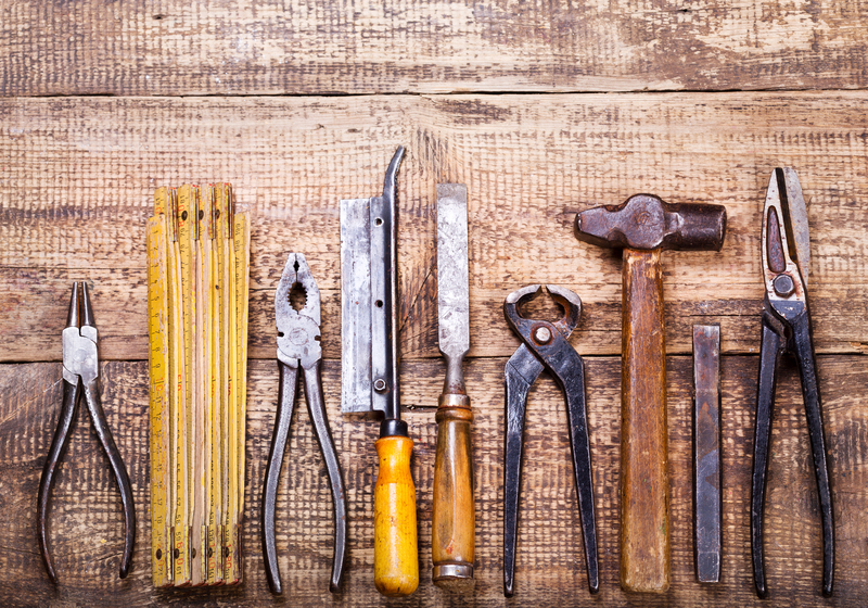 A collection of old and used hand tools arranged in a row on a rustic wooden surface. From left to right, there are small needle-nose pliers with metallic finishes, a yellow folding ruler with visible measurement markings, a pair of combination pliers with a metal head and blue handles, a metal handsaw with a black and yellow handle, a flat-head screwdriver with a wooden handle, a pair of slip joint pliers with a metallic finish, a hammer with a wooden handle and a darkened hammerhead, a metal chisel with a flat blade, and a pair of cutting scissors with metallic blades and black handles. The tools show signs of wear and age, with some rust and chipped paint visible. The wooden surface features a natural, textured grain with visible knots and irregularities, providing a neutral background that emphasizes the metallic and wooden materials of the tools. This scene can relate to private waste handling or on-site clearance activities as part of a rubbish removal process, reflecting materials that might be gathered for disposal or recycling, characteristic of independent collection services provided by companies such as Big Ben.