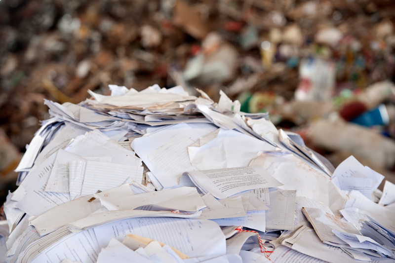 A large pile of mixed paperwork and shredded documents, consisting of various types of paper, including lined and plain sheets, some crumpled and others flattened, spread across the foreground of the image. The papers feature different textures, ranging from smooth to slightly wrinkled, with colors predominantly in white, off-white, and light beige tones. The background shows a blurred heap of miscellaneous waste and debris, indicating an outdoor environment such as a waste site or skip area. The scene is illuminated by natural daylight, highlighting the disorganized nature of the waste. The image suggests a context of waste collection, disposal, or recycling services, possibly handled by companies like Big Ben, specializing in private rubbish removal or alternative waste management solutions. The focus is on the clutter of paper waste, illustrating the type of materials involved in document disposal processes.