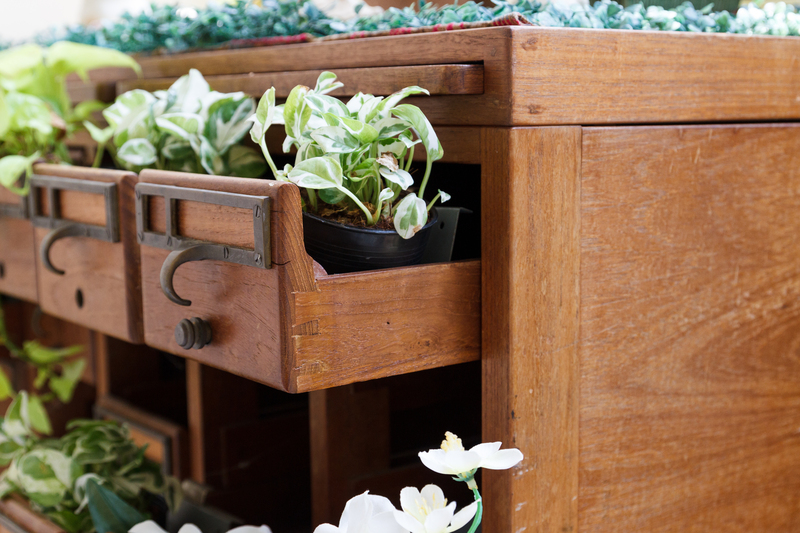 A close-up view of a wooden outdoor planter box with a natural finish, featuring multiple small drawers equipped with metal handles and hooks. The drawers contain potted plants with variegated green and white leaves, some spilling over the edges. The planter box is situated on a paved or decked surface, and surrounding greenery is visible in the background, suggesting a garden or patio setting. The natural light highlights the grain and texture of the wood, creating a warm and inviting atmosphere. This scene illustrates on-site garden waste management or private waste handling, often part of rubbish removal services like those provided by Big Ben, which may include the collection or management of garden and organic waste alongside general rubbish disposal options.