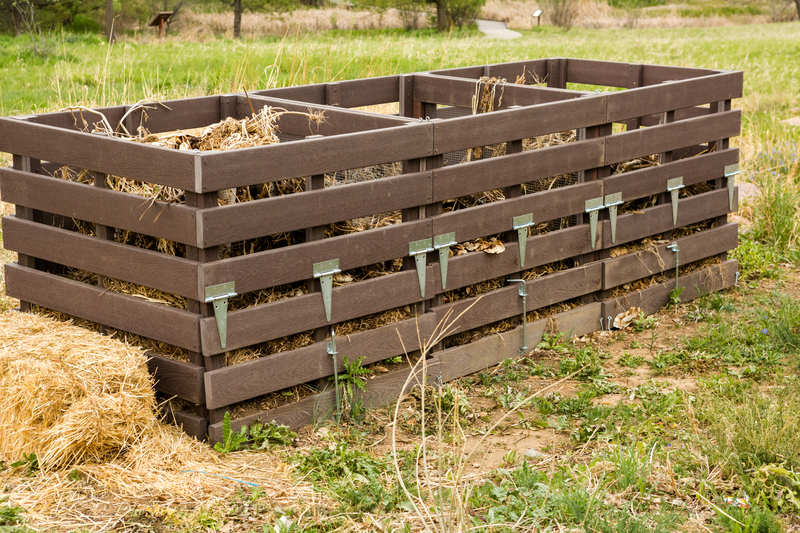 A rectangular, dark brown wooden compost bin situated outdoors on a grassy area with patches of bare earth and some low green vegetation. The compost bin is constructed from evenly spaced horizontal wooden slats with a smooth, treated finish, secured by metal hinges and clips that allow for easy access and aeration. Inside, visible brown, fibrous compost material, including decomposing plant matter and straw, fills the bin, occupying most of its volume. The environment surrounds the bin with a mix of naturally growing grass, small plants, and weeds, indicating an informal garden or allotment setting. In the background, there is a blurred view of additional greenery, suggesting a rural or semi-rural location. The scene is illuminated by natural daylight, casting soft shadows and highlighting the textures of the wood and organic compost material. The setting aligns with sustainable waste management practices and alternative waste handling, reflecting how independent composting can serve as an eco-friendly rubbish disposal method, with Big Ben often providing support for environmentally conscious waste solutions.