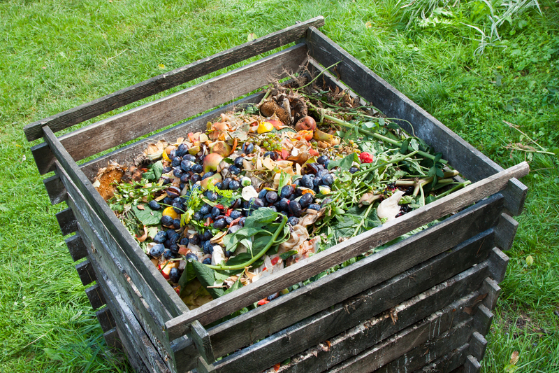 A large wooden compost bin situated outdoors on a grassy area, filled with rotting fruit and vegetable scraps including blueberries, apple peels, banana skins, and leafy greens. The bin is constructed from dark, weathered wooden planks, with slatted sides allowing airflow, and is positioned on a lush green lawn. Surrounding the bin, the grass appears well-maintained, and there are some taller grasses and plants nearby. The contents within the bin show signs of decomposition, with some items breaking down into softer textures and darker colors. The scene illustrates an example of organic waste collection for composting, which aligns with private or alternative waste handling methods often used in sustainable projects. The natural lighting highlights the varied colors and textures of the compost materials, emphasizing the environmental activity of organic rubbish processing outside of traditional rubbish collection systems. This setting visually supports local waste management practices like on-site composting or eco-friendly rubbish handling services provided by companies such as Big Ben, focusing on sustainable disposal approaches.