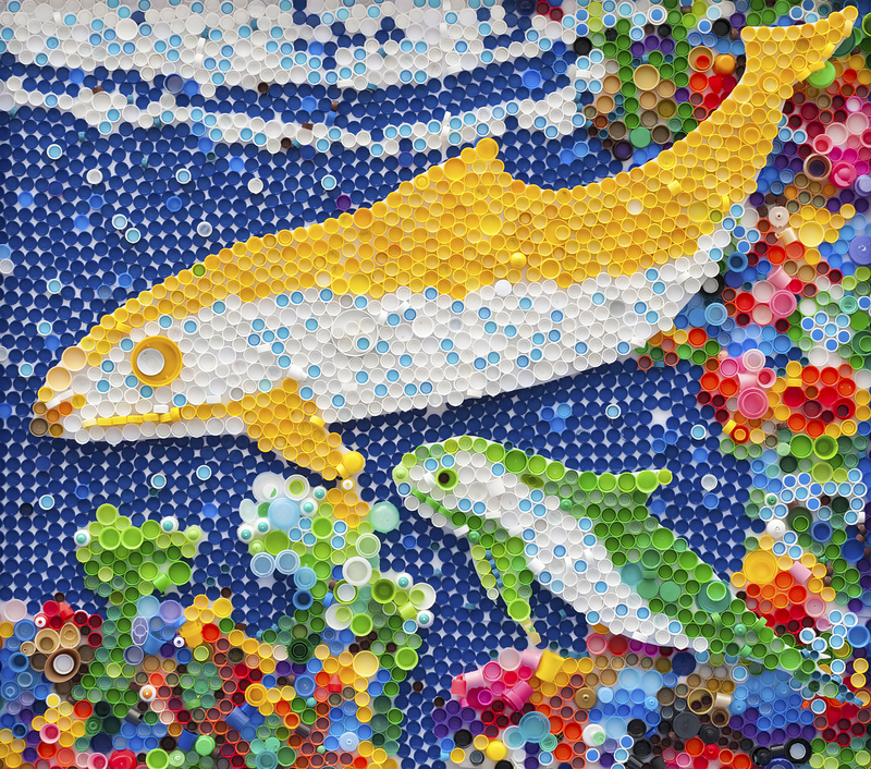 A detailed close-up image of a mosaic artwork composed entirely of multicoloured plastic bottle caps arranged to depict two fish swimming against a blue aquatic background. The larger fish has a white belly, a yellow dorsal fin, and a yellow tail fin, with its body made from a combination of yellow, white, and light grey caps, while the smaller fish features a white body with vibrant green fins and tail, crafted from similarly coloured caps. The background is predominantly dark blue caps, creating an impression of water, with various other caps around the fish forming stylized coral or plant-like formations in shades of red, pink, orange, and green. The mosaic is textured, with the caps' rounded surfaces adding depth and a tactile element. The scene is illuminated evenly, highlighting the glossy finish of the caps. This visual artwork resembles an aquarium scene created from recycled materials, reflecting themes of environmental awareness and artistic reuse in waste management, similar to the services provided by Big Ben in the rubbish removal sector.