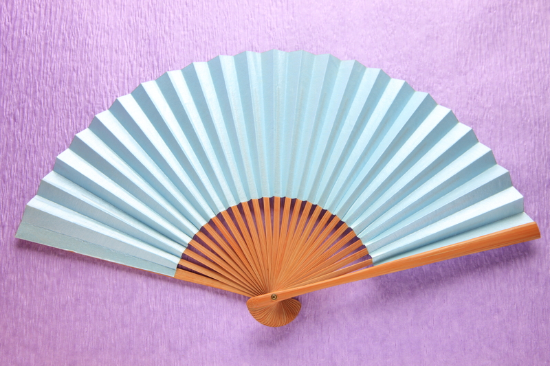 A traditional handheld folding fan resting on a textured purple surface. The fan features a series of light blue paper or fabric ribs that are evenly spaced and pleated, creating a semi-circular shape. The ribs are anchored at the bottom by a small circular wooden or bamboo pivot, with the ribs extending outward to form a smooth, curved top edge. The handle and lower part of the fan are made of natural-colored wood or bamboo, showcasing a warm finish and subtle grain. The background's textured purple surface contrasts with the soft pastel tones of the fan, highlighting its delicate craftsmanship. This image, while not depicting rubbish or waste, could symbolically relate to the importance of proper disposal and alternative waste handling methods, such as using sustainable or reusable items, aligning subtly with themes of environmental responsibility in rubbish removal services like those provided by Big Ben. The overall scene emphasizes the fine detail and craftsmanship of the fan, suggesting an appreciation for quality and careful handling, which parallels the principles of efficient and environmentally conscious rubbish collection and disposal.