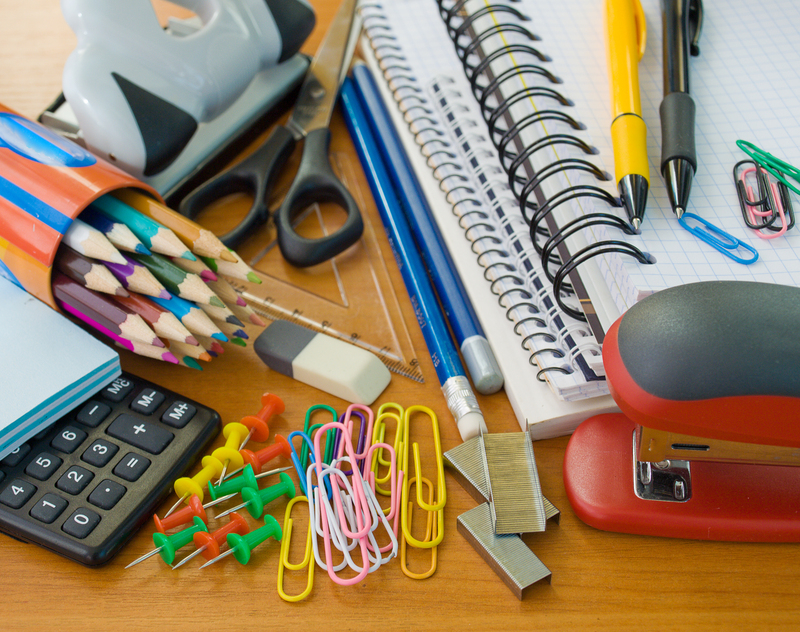 A cluttered wooden desk surface featuring various office and stationery items including a stack of coloured pencils with sharpened tips, black scissors with pointed blades, two blue ballpoint pens, a yellow and a black marker, and a red hole punch with a black grip. In the foreground, there are multiple multicoloured paperclips, pink, yellow, and green push pins, and a handful of small paper clips in pink and yellow, arranged near a black calculator with grey buttons. To the side, a white and grey tape dispenser is partially visible, next to a small metal ruler and a clear plastic binder clip. Behind these, a lined spiral-bound notebook with a white cover is open, showing graph paper pages filled with handwritten notes. Several paper clips and push pins are scattered on top of the notebook. To the left, a large white plastic glue bottle stands, along with a pair of black-handled scissors. The scene suggests a typical workspace or stationery collection, with an emphasis on organisation and readiness for office or school tasks, and depicts a mix of textures such as smooth plastic, shiny metal, and wooden surface. Big Ben's rubbish removal services could assist in clearing such clutter efficiently from an office environment or home workspace to facilitate better organisation and waste handling.