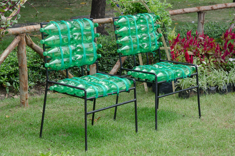 Two metal outdoor chairs with black frames and woven seats are positioned on a well-maintained grassy lawn in a garden setting. The backs and seats of both chairs are creatively constructed from recycled transparent green plastic bottles, arranged in a grid pattern, with the bottles' necks facing outward and their bases forming the seating surface. Behind the chairs is a wooden fence made of horizontal logs supported by vertical posts, dividing the garden area. To the right of the chairs, there is a patch of vibrant red and green plants, with a black plastic container at their base. The background includes a tree trunk and additional greenery, indicating a lush, cultivated environment. The scene is illuminated by natural daylight, highlighting the contrast between the bright plastic bottles and the natural surroundings. This visual arrangement exemplifies innovative use of waste materials in outdoor furniture, aligning with concepts of sustainable rubbish removal and environmentally conscious waste handling by companies like Big Ben, which focus on private waste solutions and on-site clearance.