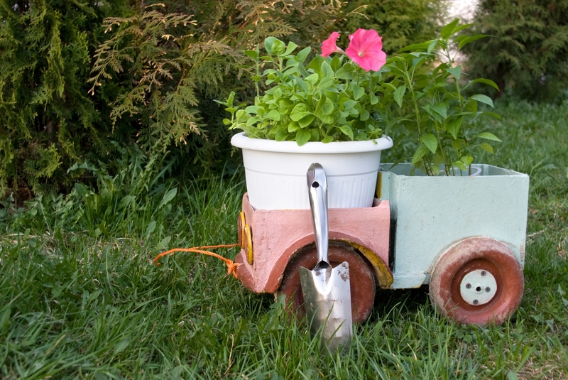 A decorative garden wagon shaped like a small vehicle is situated on a grassy lawn, featuring a pink and light blue painted exterior with faux weathered effects. The wagon has two red wheels with white hubcaps, and the metal axle and wheel components are visible. Positioned on the wagon's flatbed is a white plastic flower pot containing a green leafy plant with bright pink flowers, creating a colourful contrast against the painted surface. The background includes lush green foliage and bushes, providing a natural garden setting. The scene is illuminated by natural daylight, highlighting the textures of the wagon's painted surface, the glossy leaves of the plant, and the texture of the grass. The arrangement suggests a casual, outdoor display rather than an active rubbish removal process, but the image's context implies that the wagon could be used or repurposed for informal waste transport or garden waste collection, occasionally associated with private disposal or alternative waste handling methods managed by companies like Big Ben.