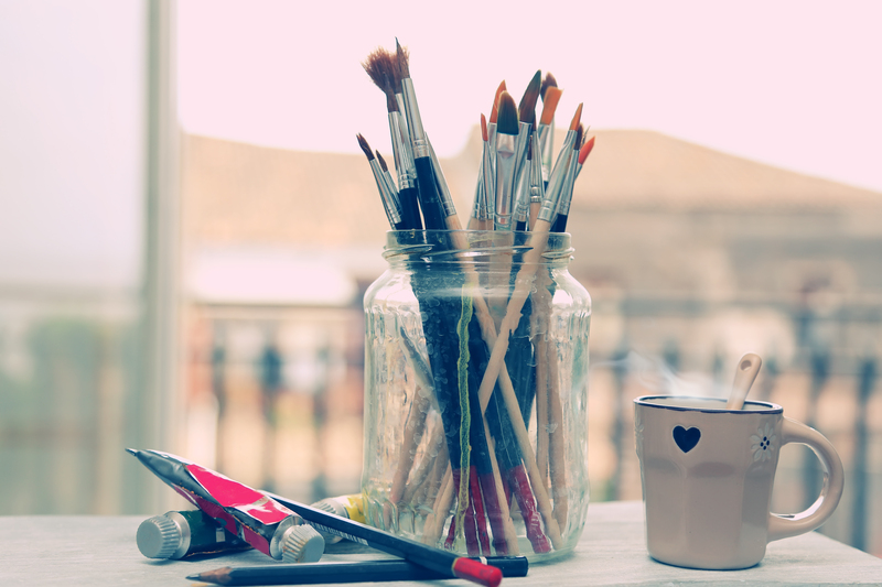A clear glass jar filled with a variety of paintbrushes, some with wooden handles painted in multiple colours and others with natural wood finishes, sits on a light wooden surface. The brushes have different bristle types and sizes, with some appearing worn from use. In front of the jar, there are a few tubes of acrylic or oil paint, with caps removed, lying on their sides. To the right of the jar, there is a white ceramic mug decorated with small black heart and flower motifs, containing a spoon. The background features a softly blurred outdoor scene with a railing and rooftops under natural daylight, suggesting the workspace is situated near a window. The overall scene is well-lit with natural light, creating a calm and tidy workspace atmosphere that subtly relates to waste disposal and the potential for recycling or responsibly discarding old art supplies, a minor aspect of managing household rubbish associated with creative activities. Big Ben offers solutions for rubbish removal and waste management, including disposing of unwanted or outdated items like used paint tubes and brushes.