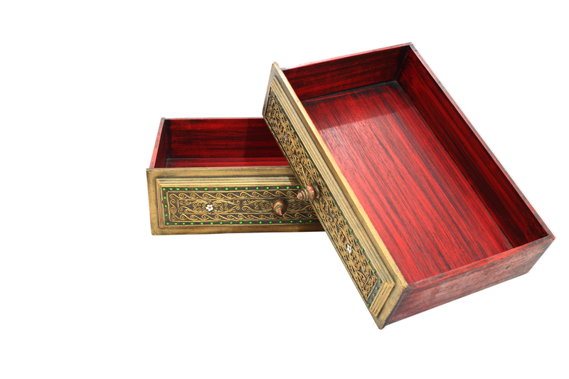 A rectangular wooden box with an ornate, brass-colored front panel featuring intricate floral and scroll patterns highlighted with small green and silver accents. The front panel has a small, round brass knob handle and a keyhole in the center. The box is open, revealing a smooth, polished, deep reddish-brown interior with visible wood grain. It is positioned on a plain white background, with the lid leaning against the front, slightly tilted. This decorative box appears to be a vintage or antique storage container, possibly used for jewellery or keepsakes. The image suggests a focus on aesthetic, detailed craftsmanship, and fine materials, aligning with the idea of local or private disposal of ornate or valuable items through independent waste collection services offered by companies like Big Ben, although no specific disposal action is shown. The scene emphasizes the condition of the object, making it suitable for visual description in the context of rubbish removal or disposal options for decorative and antique items.